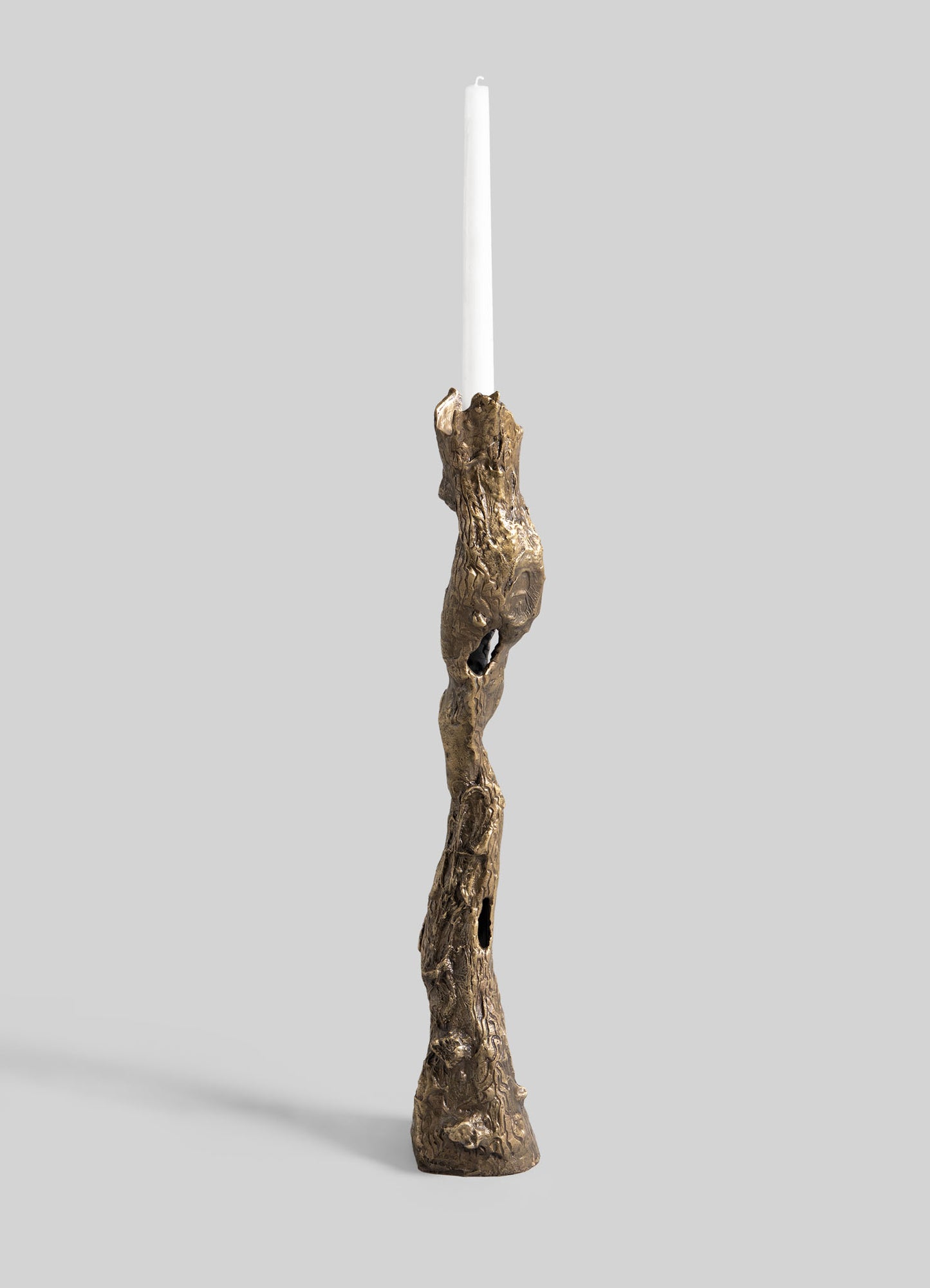 A tall white candle sits in the VIYA Kikar Candle Holder Large, a sculptural brass piece with twisted, branch-like forms that highlight timeless metalwork, set against a plain light gray background.