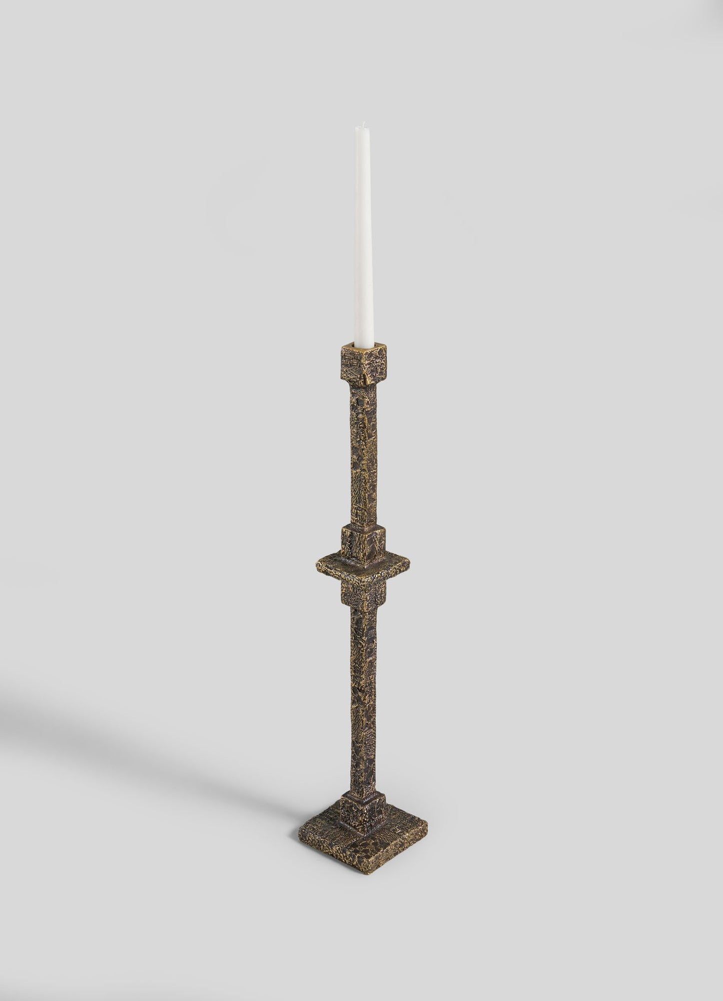 The VIYA Tiruppur Totem Candle Holder Small, a tall ornate brass piece with a white taper candle, square base, and brocade-inspired detailing, is displayed against a light gray background.