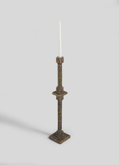 The VIYA Tiruppur Totem Candle Holder Small, a tall ornate brass piece with a white taper candle, square base, and brocade-inspired detailing, is displayed against a light gray background.