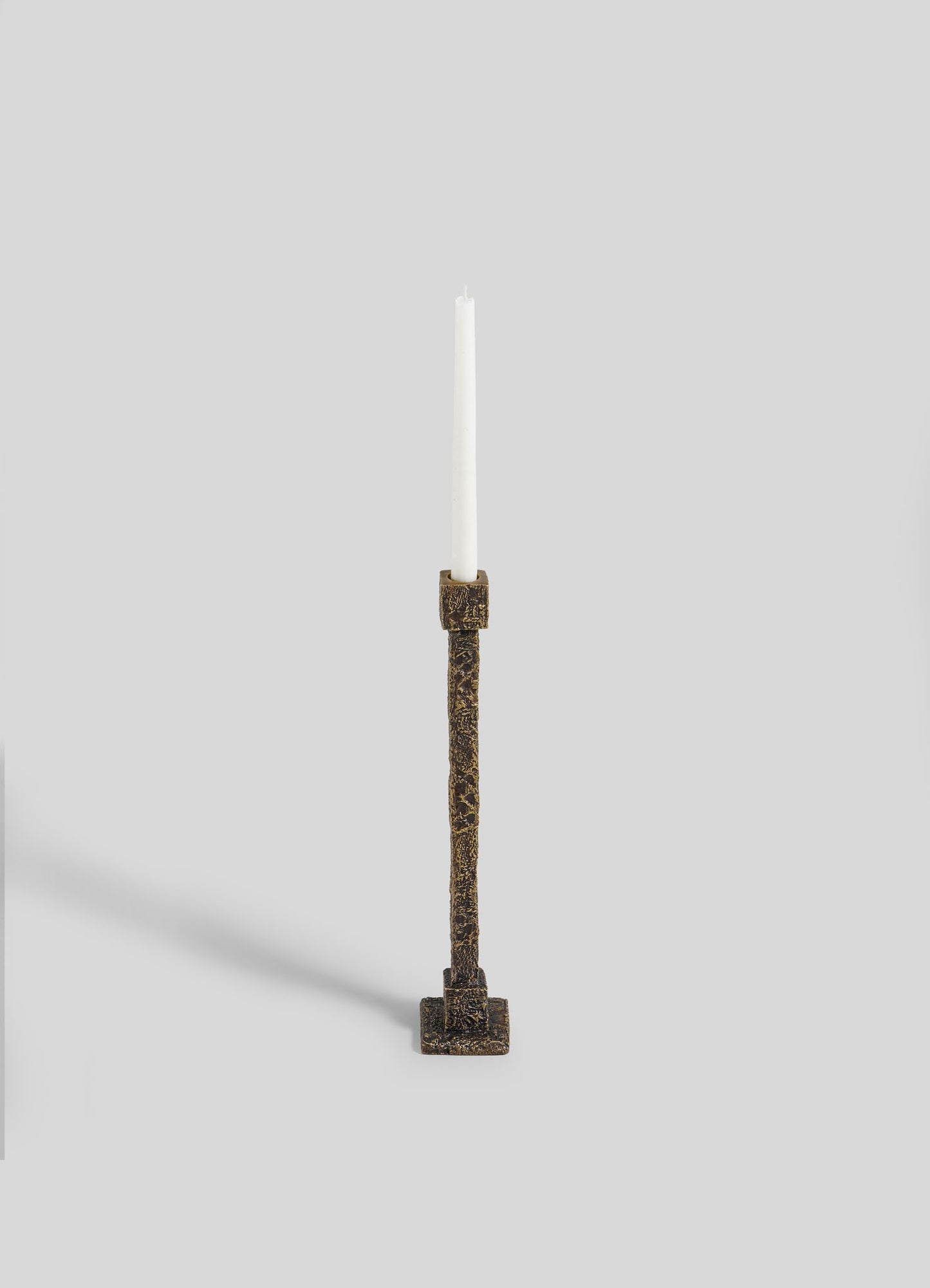 The VIYA Tiruppur Candle Holder Large, featuring ornate textured bronze with a square base inspired by Indian textiles, displays a tall white candle. Set against a plain light gray background, the candle casts a faint shadow to the left.