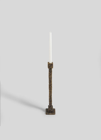 The VIYA Tiruppur Candle Holder Large, featuring ornate textured bronze with a square base inspired by Indian textiles, displays a tall white candle. Set against a plain light gray background, the candle casts a faint shadow to the left.
