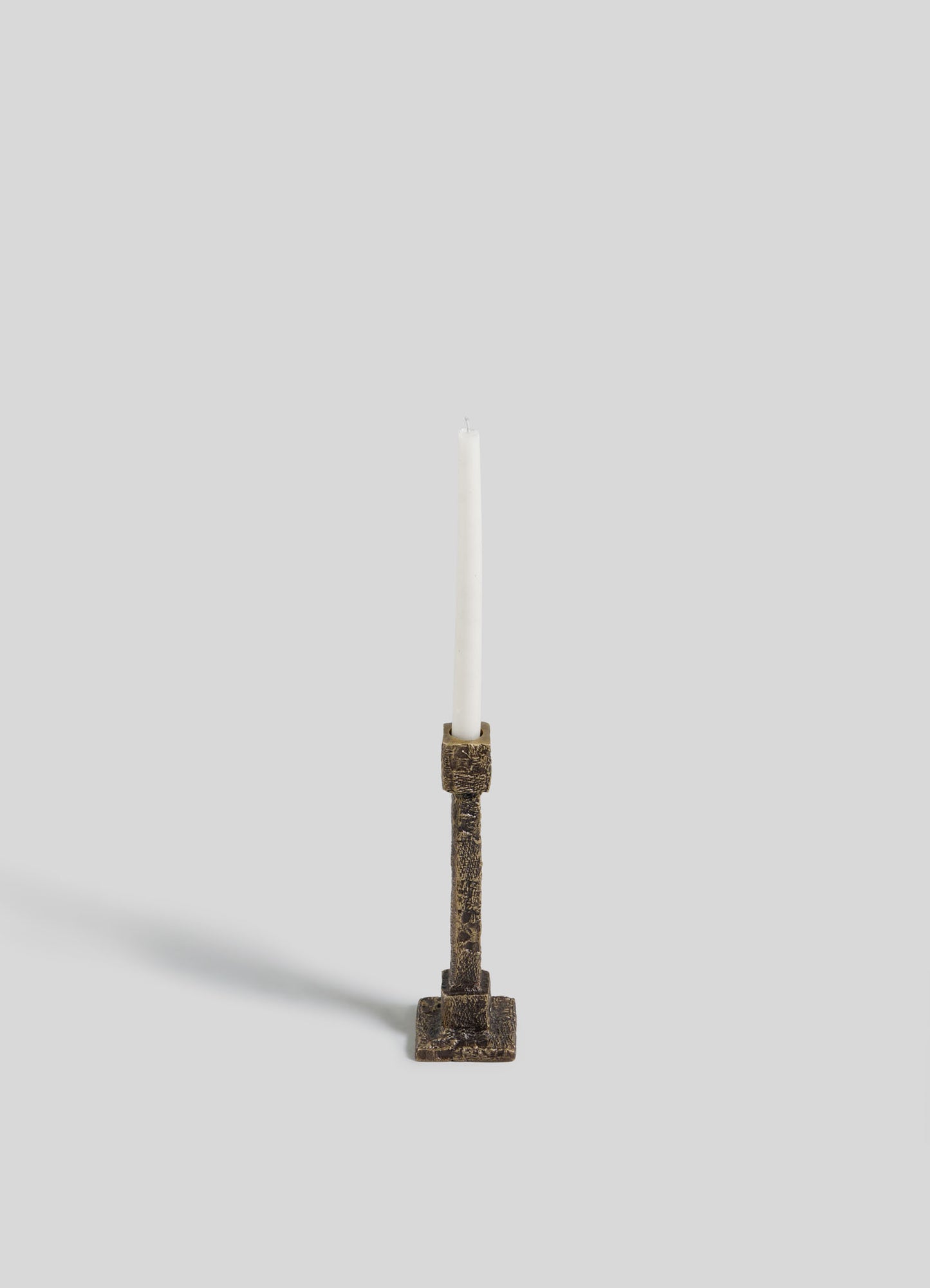 The VIYA Tiruppur Candle Holder Small, with an antique-style bronze finish reminiscent of Indian brocades, holds a single white candle upright against a plain light gray background. The candle’s soft shadow falls to the left.