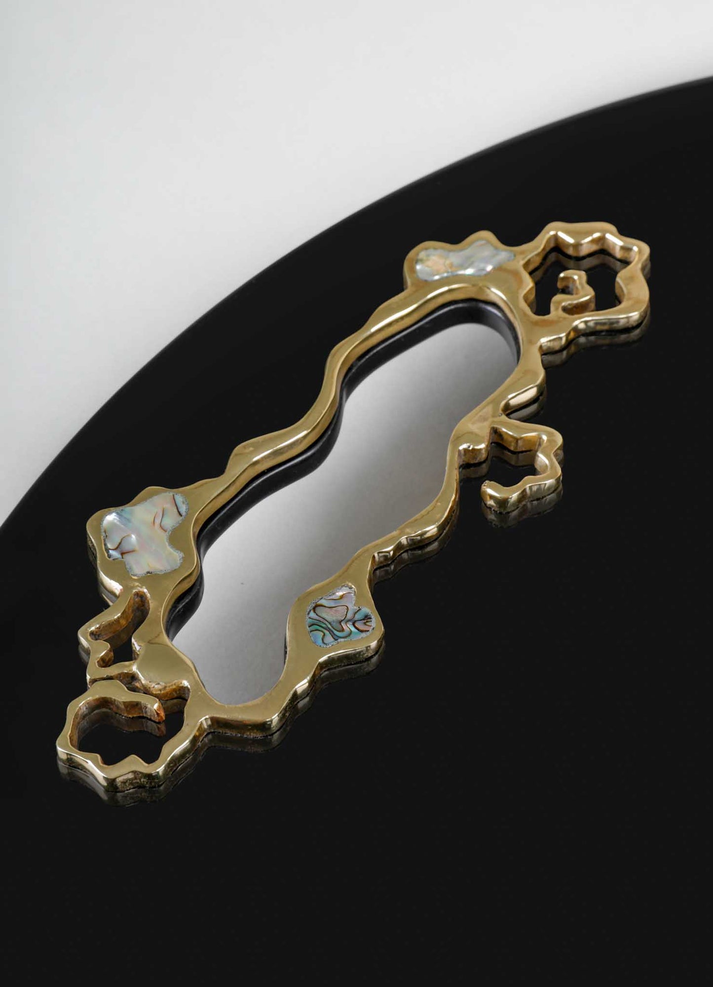 A gold-toned jewelry pendant with an irregular, organic shape featuring inlaid mother-of-pearl accents, displayed on a black reflective surface.
