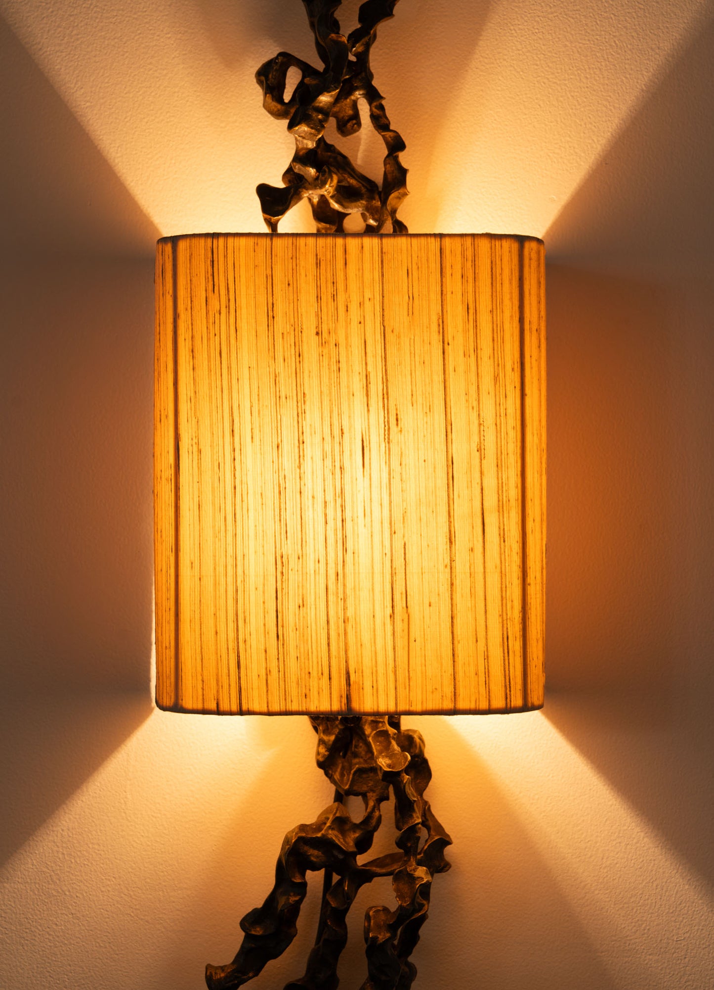 The Andaman Wall Sconce A by VIYA features a cylindrical, textured shade that casts warm light up and down, creating unique wall patterns. Decorative twisted metal elements inspired by organic forms accent the vertical back of the lamp.