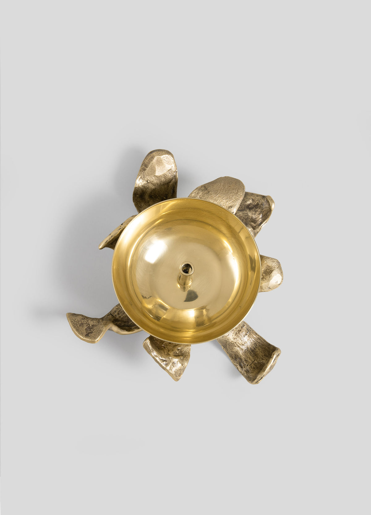 A top view of the Glory Lily Eternal Diya by VIYA shows a smooth, round gold metal bowl resting on an abstract, textured gold base with organic, curved shapes, set against a plain gray background—an elegant piece that channels ancient metalworking techniques into a modern heirloom.