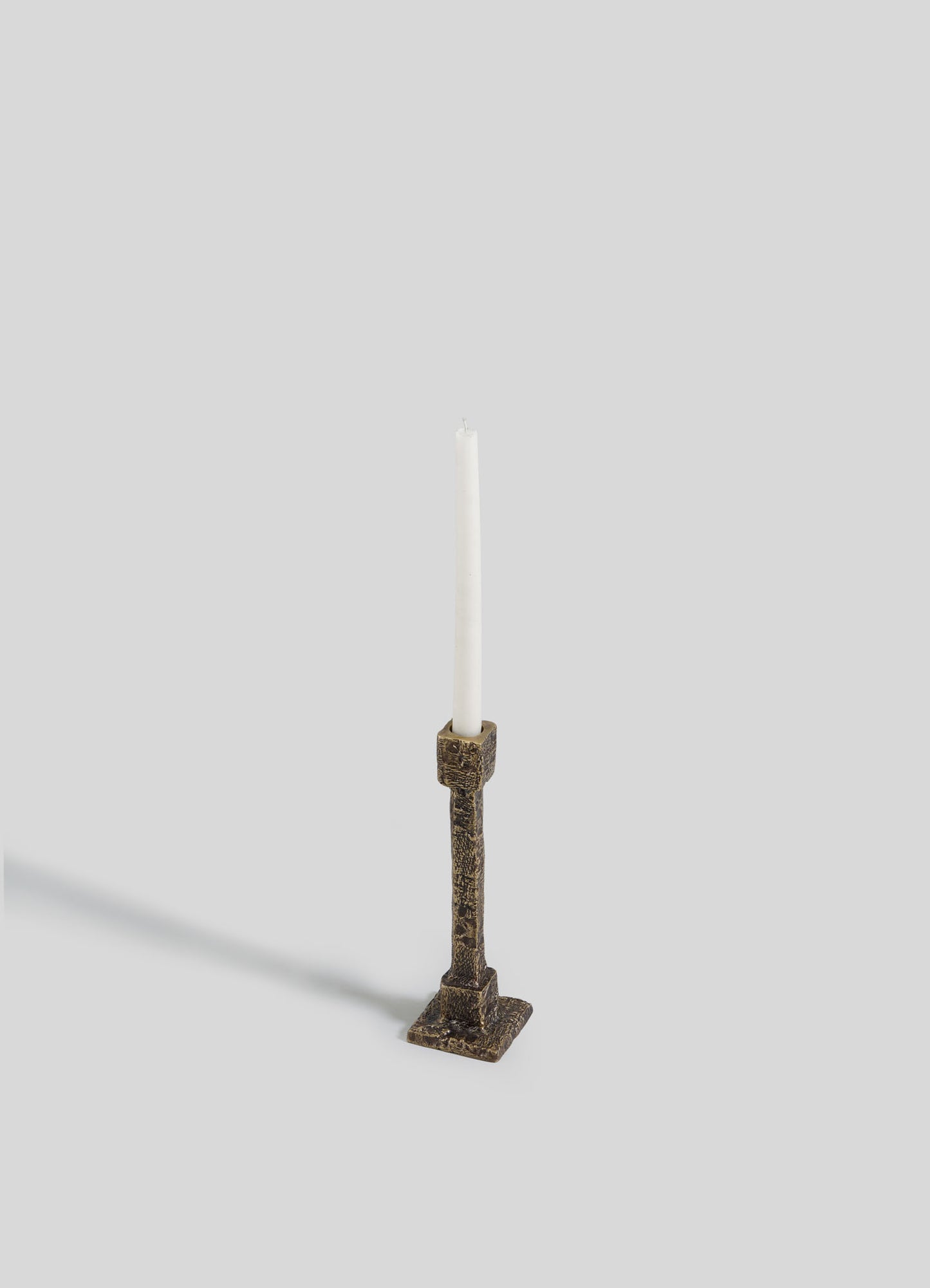 A single white taper candle stands upright in the VIYA Tiruppur Candle Holder Small, an ornate brass piece inspired by Indian brocades and metal textiles, set against a light gray background with a soft shadow cast to the left.
