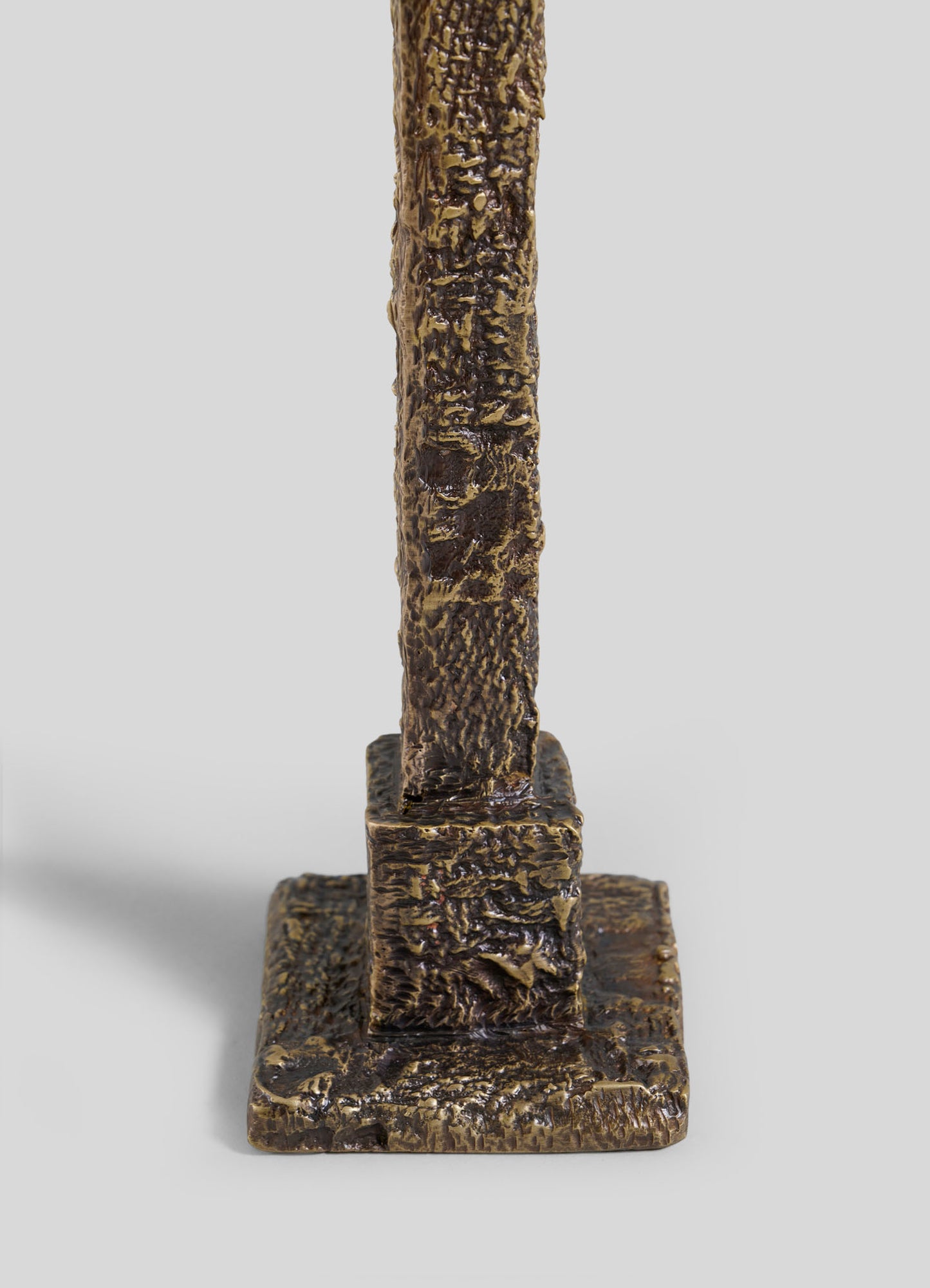 A close-up of the VIYA Tiruppur Candle Holder Large, a textured bronze-colored vertical sculpture on a rectangular base, its surface inspired by intricate Indian textile patterns, shown against a plain gray background.