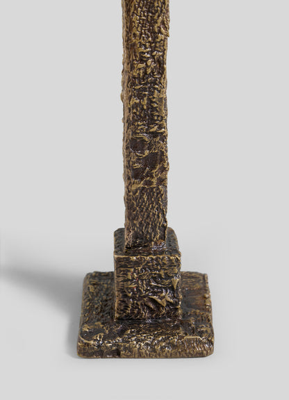 A close-up of the VIYA Tiruppur Candle Holder Large, a textured bronze-colored vertical sculpture on a rectangular base, its surface inspired by intricate Indian textile patterns, shown against a plain gray background.