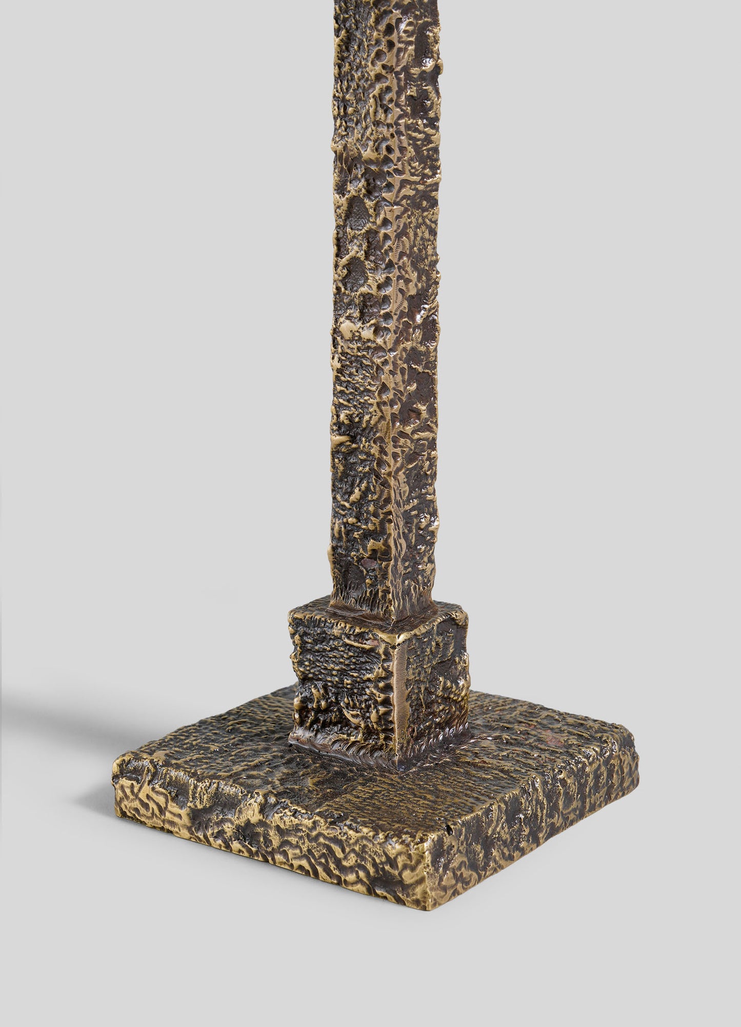 A close-up of the Tiruppur Totem Candle Holder Small by VIYA, featuring a bronze-toned, sculpted metal form with intricate patterns and a textured, rough surface, reminiscent of metal textiles on its square base.