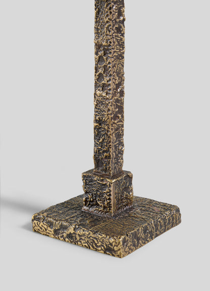 A close-up of the Tiruppur Totem Candle Holder Small by VIYA, featuring a bronze-toned, sculpted metal form with intricate patterns and a textured, rough surface, reminiscent of metal textiles on its square base.