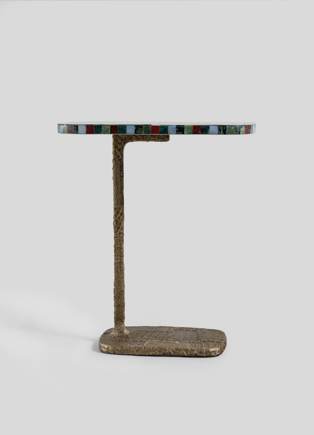 The VIYA Mosaic Red Side Table features a round tabletop with a multicolored, mosaic-inspired edge and is supported by a textured, bronze-hued L-shaped cast brass base. The table is displayed against a plain light gray background.