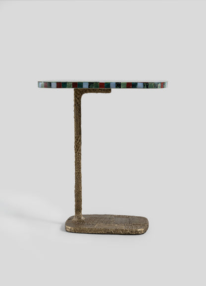 The VIYA Mosaic Red Side Table features a round tabletop with a multicolored, mosaic-inspired edge and is supported by a textured, bronze-hued L-shaped cast brass base. The table is displayed against a plain light gray background.