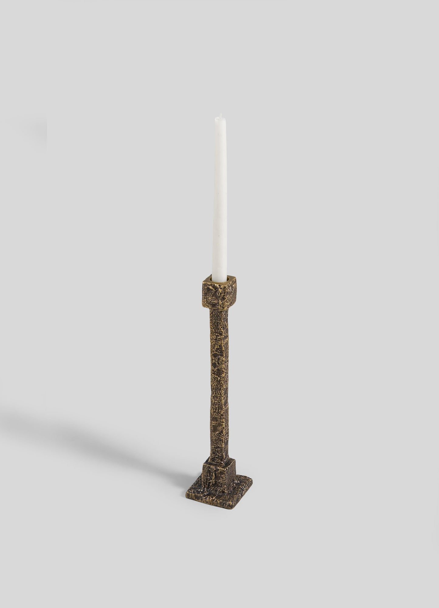The VIYA Tiruppur Candle Holder Medium features a tall, textured bronze design with a square base, holding a single white taper candle, all set against a plain light gray background.