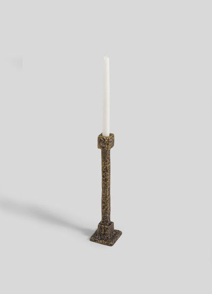 The VIYA Tiruppur Candle Holder Medium features a tall, textured bronze design with a square base, holding a single white taper candle, all set against a plain light gray background.