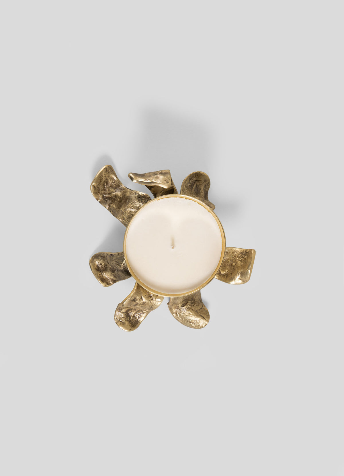 The Glory Lily Scented Diya by Viya features a white candle resting in a round, gold-toned holder with six textured, curved petal-like shapes reminiscent of ancient metalworking, showcased against a plain gray background.