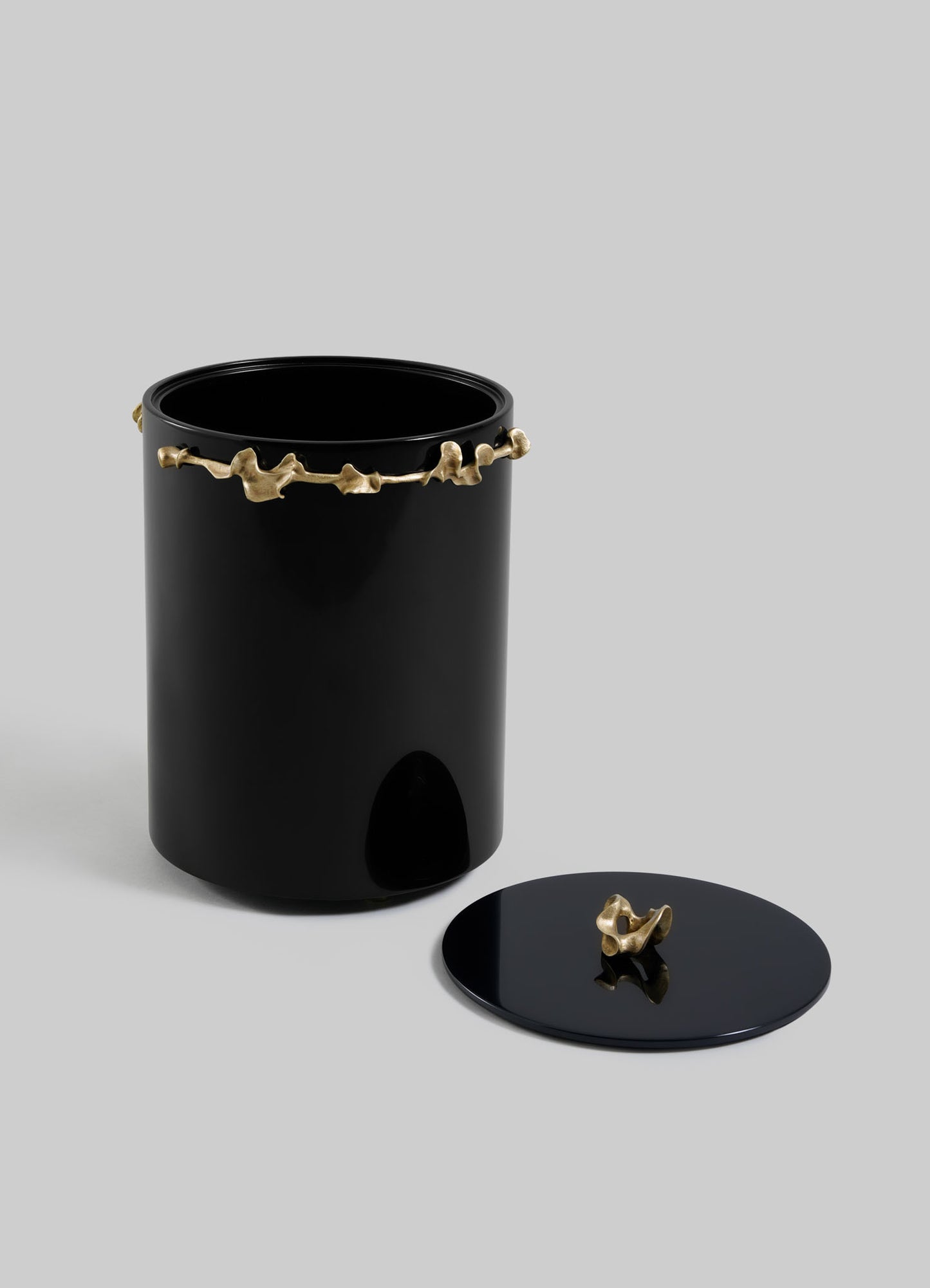 The VIYA Saffron Black Bin With Lid features a glossy black cylindrical design with a matching lid, enhanced by a gold branch detail and handle, creating an elegant look against a plain gray background.