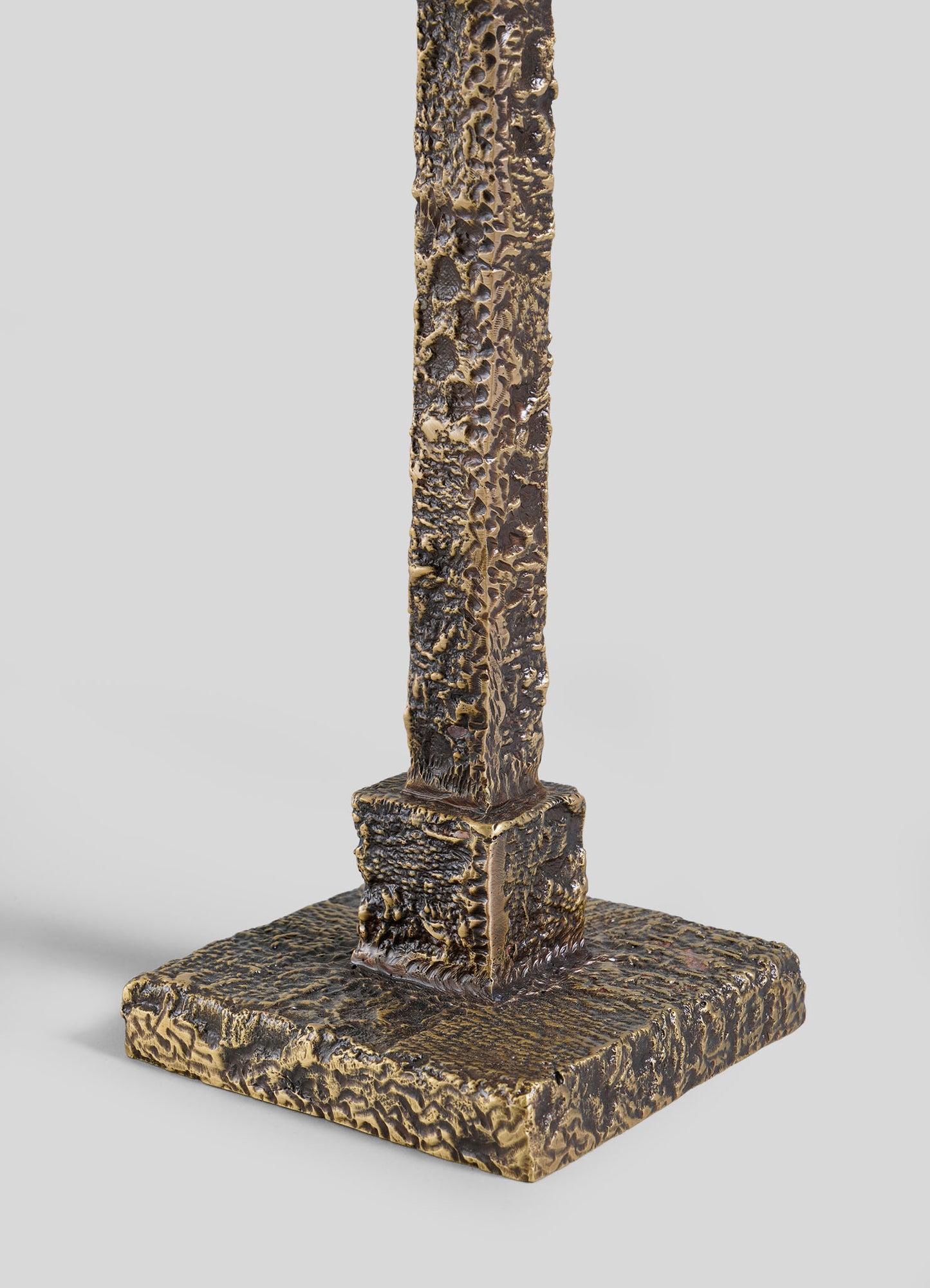 The VIYA Tiruppur Totem Candle Holder Large features a textured, bronze-colored metal post on a square base, its rough surface evoking brocade patterns, set against a plain light gray background.