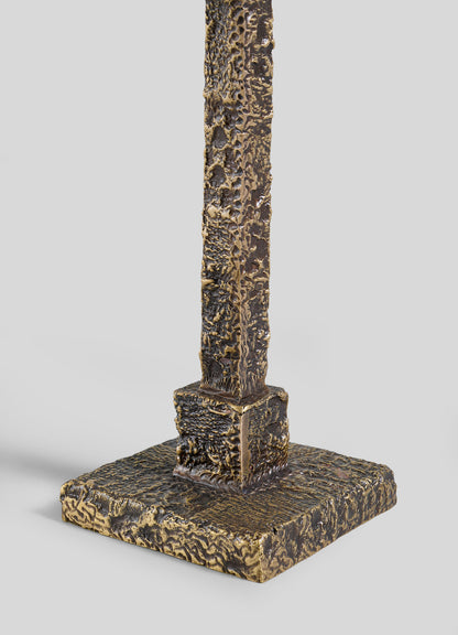 The VIYA Tiruppur Totem Candle Holder Large features a textured, bronze-colored metal post on a square base, its rough surface evoking brocade patterns, set against a plain light gray background.