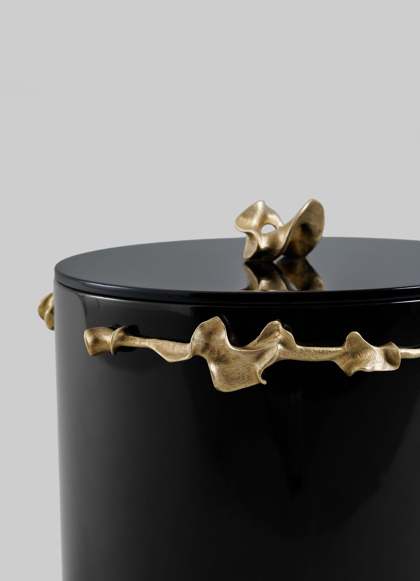The VIYA Saffron Black Bin With Lid is a glossy black cylindrical container, accented with an abstract gold metallic ribbon and a matching sculptural handle on top, shown against a plain gray background.