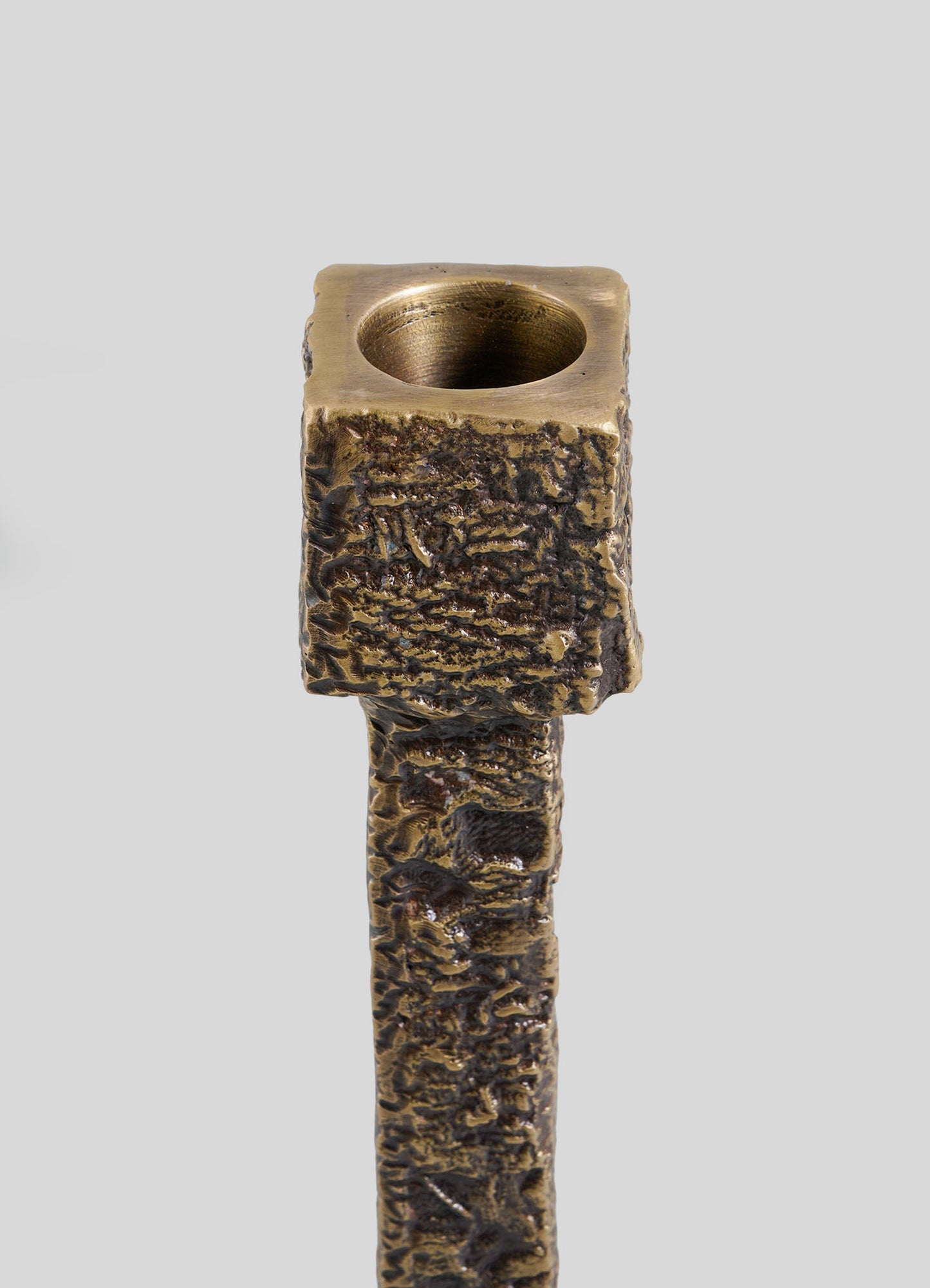 A close-up of the VIYA Tiruppur Candle Holder Large, featuring a textured bronze finish with a square top and round candle holder opening inspired by Indian textiles, set against a plain grey background.