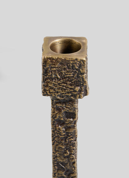 A close-up of the VIYA Tiruppur Candle Holder Large, featuring a textured bronze finish with a square top and round candle holder opening inspired by Indian textiles, set against a plain grey background.