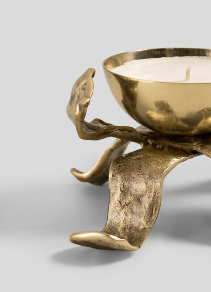 A close-up of the Glory Lily Scented Diya by Viya showcases its golden, branch-inspired legs cradling a round white candle. Crafted with an abstract and textured design reminiscent of ancient metalworking techniques, this unique candle holder stands out against a plain grey background.