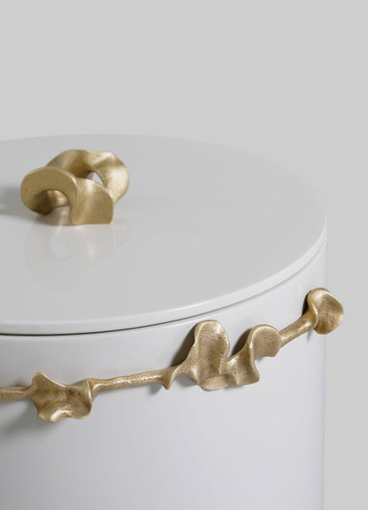 The VIYA Saffron Ecru Bin With Lid is a white cylindrical container with a smooth surface, highlighted by a sculptural gold handle and an abstract gold accent wrapping around the upper rim, set against a light grey background.
