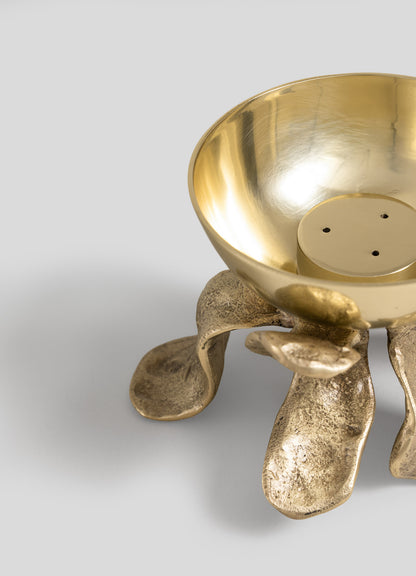 The Glory Lily Incense Diya by VIYA features a gold metal bowl with a round interior, crafted using ancient metalworking techniques, and rests atop sculpted, textured legs resembling organic forms, set against a light gray background.