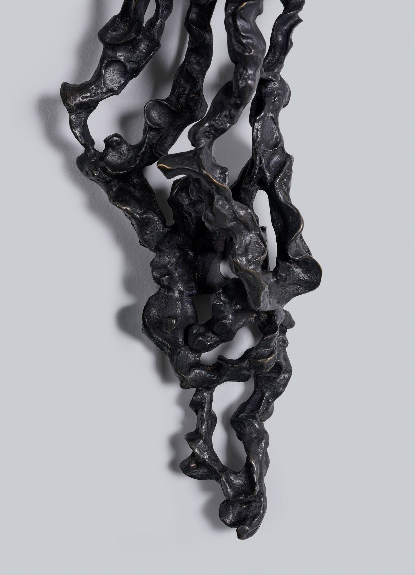The Andaman Wall Sconce A by VIYA features a twisted black sculptural form with organic curves and hollow sections, mounted vertically on a light gray background. Shadows highlight its intricate, irregular surface texture.