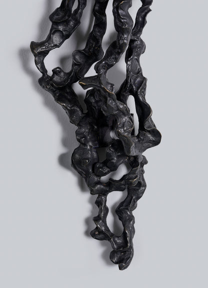 The Andaman Wall Sconce A by VIYA features a twisted black sculptural form with organic curves and hollow sections, mounted vertically on a light gray background. Shadows highlight its intricate, irregular surface texture.