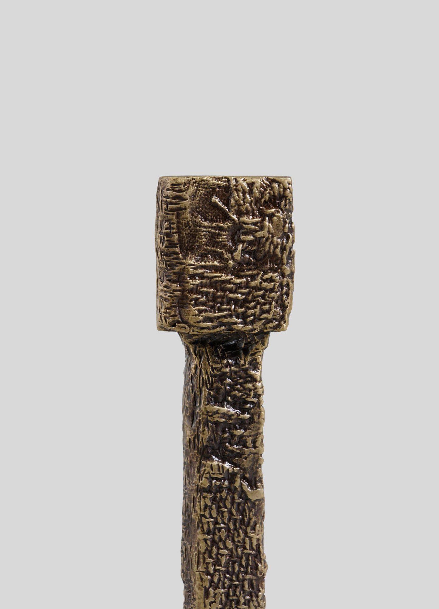 The VIYA Tiruppur Candle Holder Small features a textured bronze or brass finish with a square holder and intricate patterns, echoing Indian brocade designs, set against a plain gray backdrop.