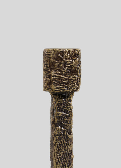 The VIYA Tiruppur Candle Holder Small features a textured bronze or brass finish with a square holder and intricate patterns, echoing Indian brocade designs, set against a plain gray backdrop.