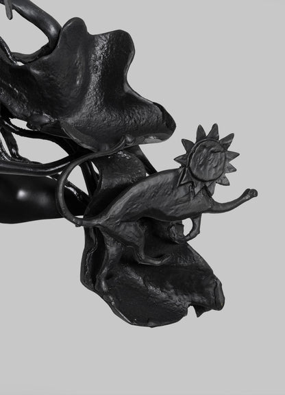 The VIYA Gardens Of India Pendant Light features a black metal dinosaur-like figure with a sun-shaped head standing on textured leaves, reflecting Indian garden motifs, set against a plain gray background.