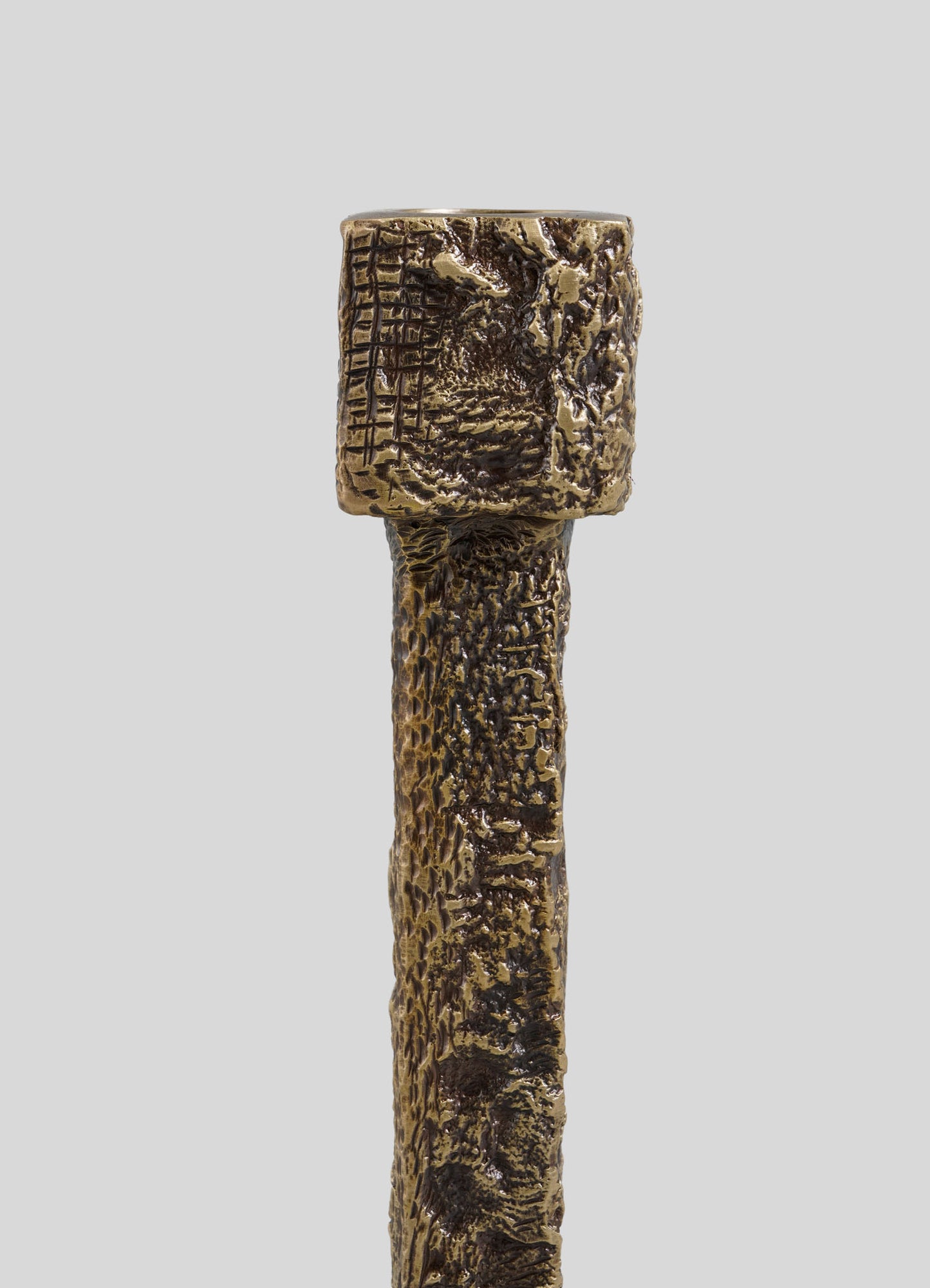 A close-up of the VIYA Tiruppur Candle Holder Medium—a textured, cylindrical bronze piece with a rough surface and irregular top—showcases intricate brocade-like patterns against a plain gray background.