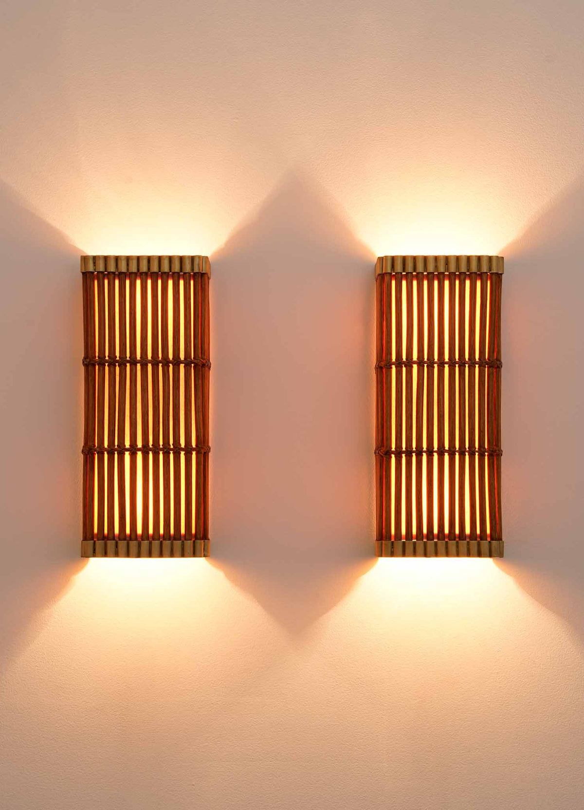 Two Namsai Wall Sconce lights by VIYA, mounted on a plain, light-colored wall, emit a warm and soft glow that creates a cozy ambiance. Meticulously handcrafted using traditional Indian techniques, these bamboo cane sconces blend artisanal beauty with inviting illumination.