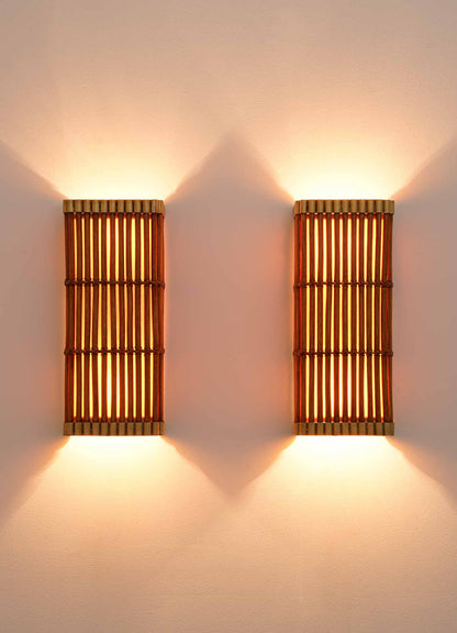 Two Namsai Wall Sconce lights by VIYA, mounted on a plain, light-colored wall, emit a warm and soft glow that creates a cozy ambiance. Meticulously handcrafted using traditional Indian techniques, these bamboo cane sconces blend artisanal beauty with inviting illumination.