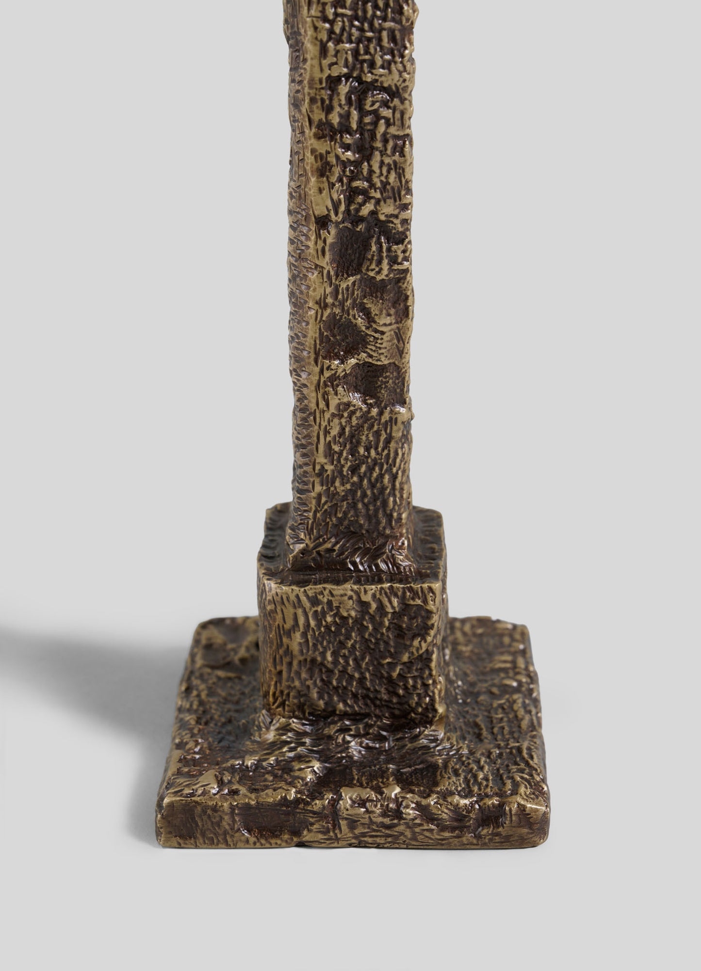 A close-up of the VIYA Tiruppur Candle Holder Small showcases its textured bronze base with a rough, textile-like surface on a square platform, set against a plain light gray background.