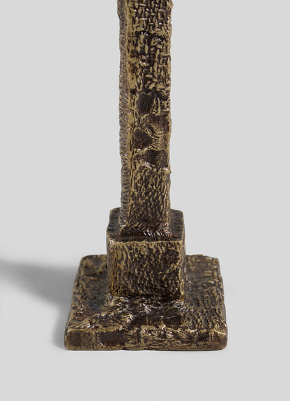 A close-up of the VIYA Tiruppur Candle Holder Small showcases its textured bronze base with a rough, textile-like surface on a square platform, set against a plain light gray background.