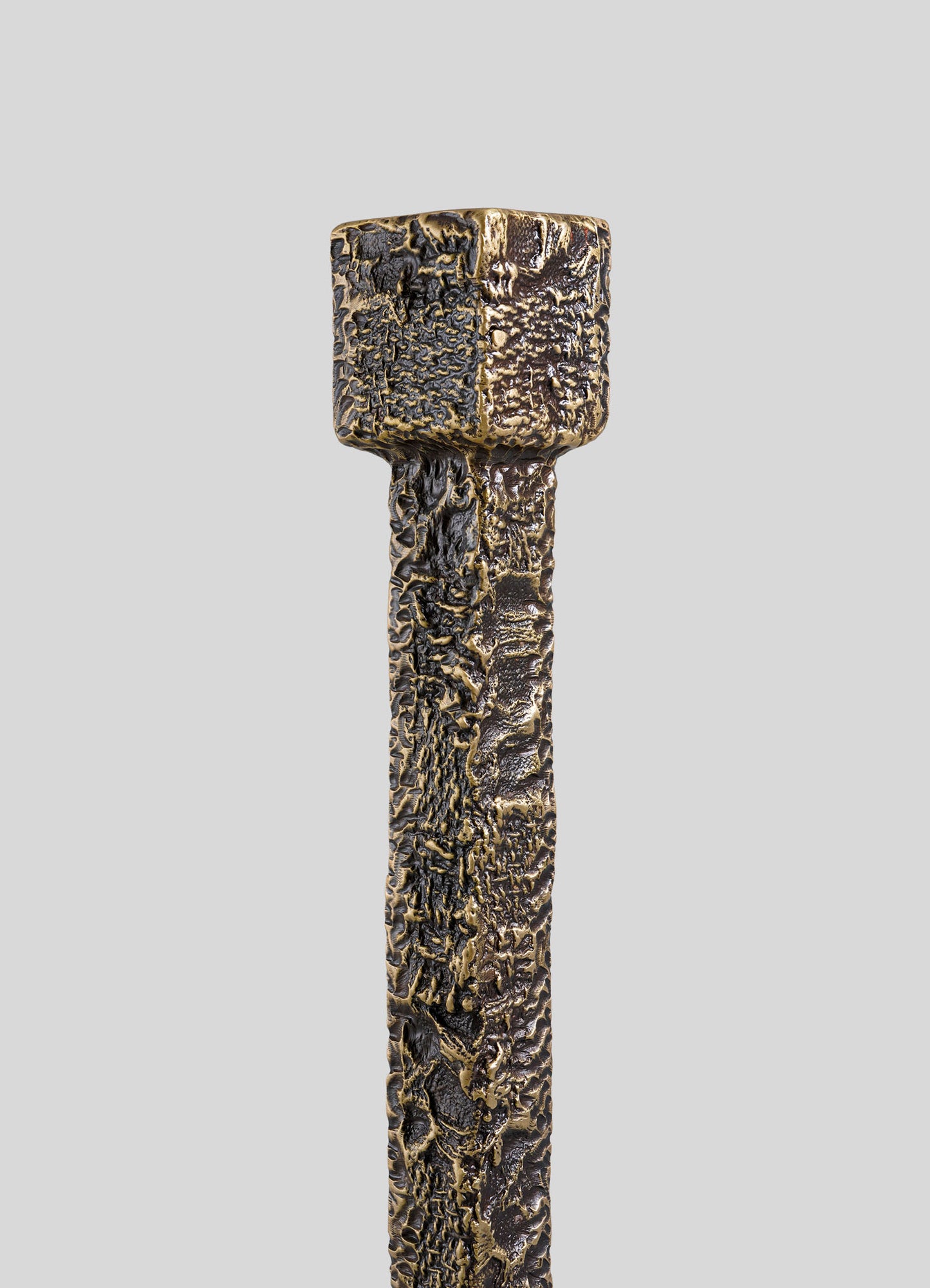A close-up of the VIYA Tiruppur Totem Candle Holder Small highlights its textured, square-shaped metallic top and rich bronze tones, showcasing its intricate metal textile-inspired design against a plain gray background.