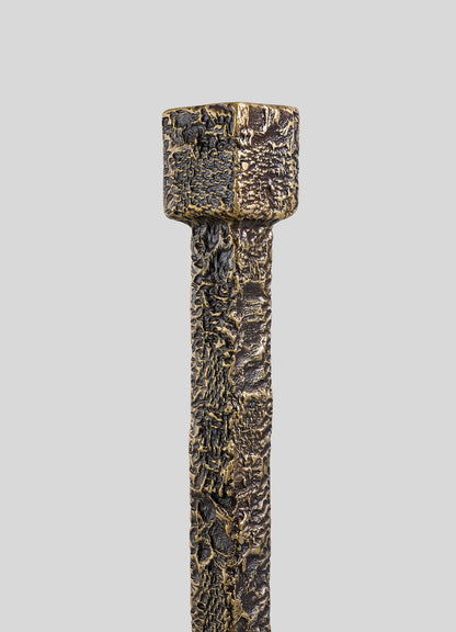 A close-up of the VIYA Tiruppur Totem Candle Holder Small highlights its textured, square-shaped metallic top and rich bronze tones, showcasing its intricate metal textile-inspired design against a plain gray background.