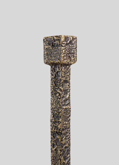 A close-up of the VIYA Tiruppur Totem Candle Holder Large, featuring a textured, metallic finish with a square top and cylindrical body reminiscent of handwoven fabrics, set against a plain light gray background.