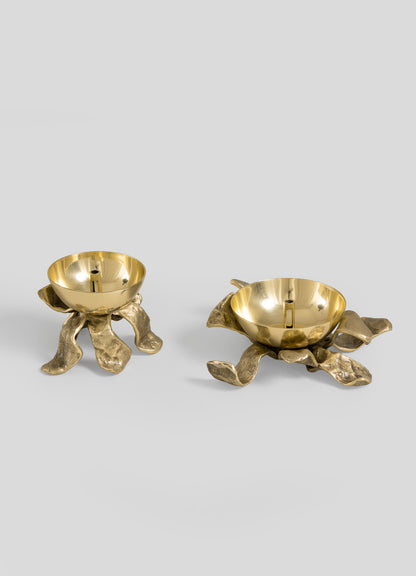 Two lustrous Glory Lily Eternal Diyas by VIYA rest atop sculptural bronze bases inspired by abstract, organic forms, their design recalling ancient metalworking artistry. Set against a plain light grey background, these pieces evoke the sense of a modern heirloom destined to be cherished for generations.