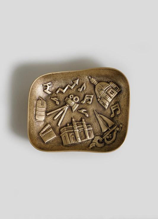 The VIYA Bombay Trinket Bowl is a decorative brass dish featuring raised icons—a film camera, musical notes, sailboat, cell phone, currency symbols, dome building, bridge, and arrow—set against a neutral background.