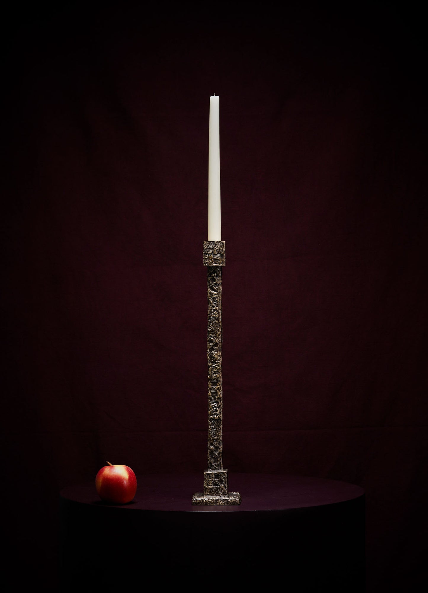 The VIYA Tiruppur Candle Holder Large, featuring an unlit white candle, stands on a dark surface beside a red apple, set against a deep burgundy background inspired by rich Indian textiles and elegant brocades.