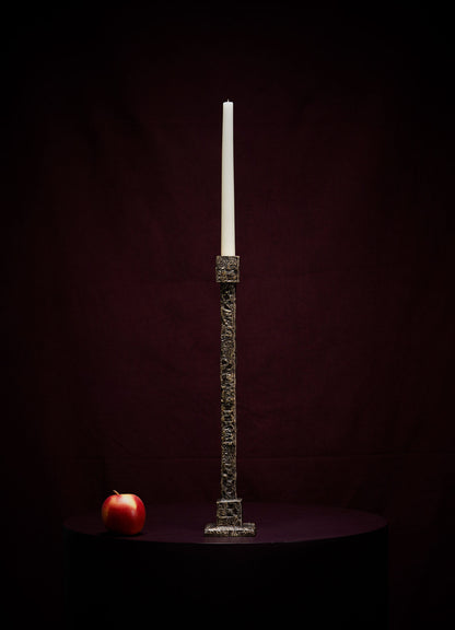 The VIYA Tiruppur Candle Holder Large, featuring an unlit white candle, stands on a dark surface beside a red apple, set against a deep burgundy background inspired by rich Indian textiles and elegant brocades.