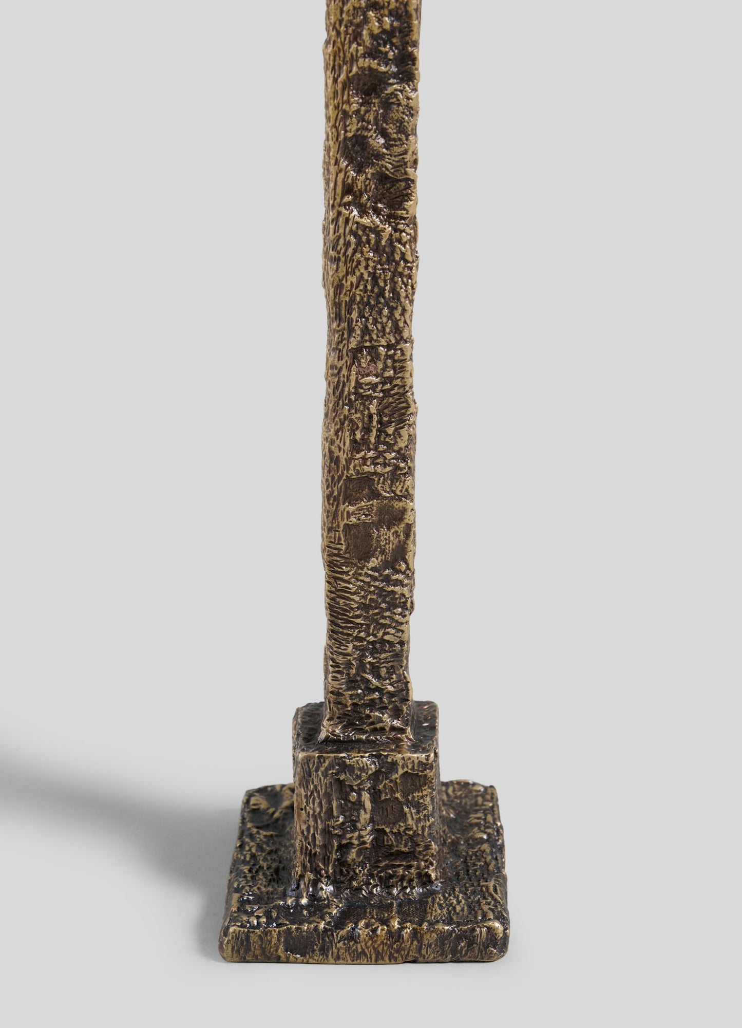 A close-up of the VIYA Tiruppur Candle Holder Medium shows its textured vertical bronze design, featuring a rough brocade-like surface on a small rectangular base, set against a plain light gray background.