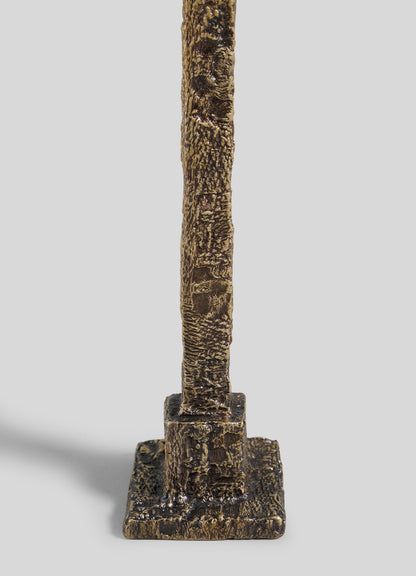 A close-up of the VIYA Tiruppur Candle Holder Medium shows its textured vertical bronze design, featuring a rough brocade-like surface on a small rectangular base, set against a plain light gray background.