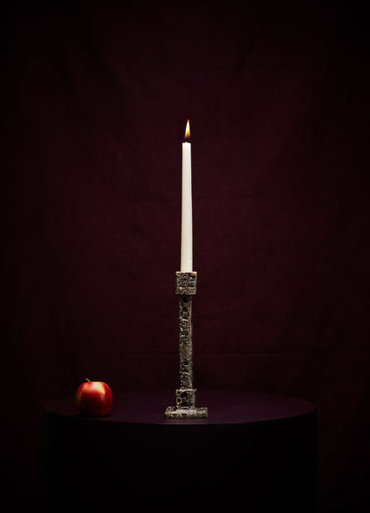 A single white candle in the VIYA Tiruppur Candle Holder Small sits on a dark round table, with a red apple to the left, set against a deep burgundy background evoking luxurious Indian brocades and handwoven fabrics.