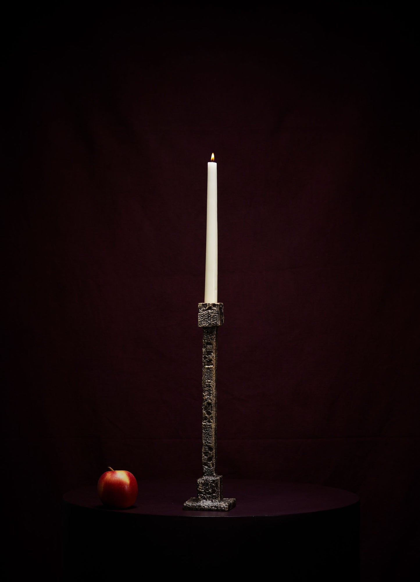 The VIYA Tiruppur Candle Holder Medium, featuring a tall white candle, stands on a dark round surface with a red apple to its left, set against a rich maroon brocade-like background.