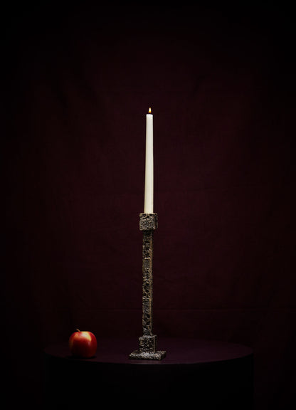 The VIYA Tiruppur Candle Holder Medium, featuring a tall white candle, stands on a dark round surface with a red apple to its left, set against a rich maroon brocade-like background.